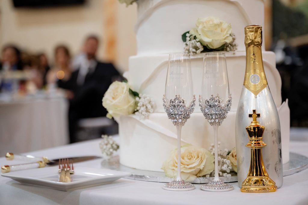 Crystal-trimmed champagne flutes and a gold-topped bottle beside a white rose wedding cake