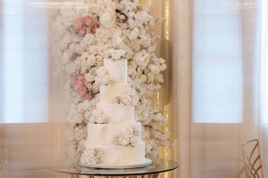 Tiered white wedding cake with ivory roses and baby's breath in front of a floral backdrop